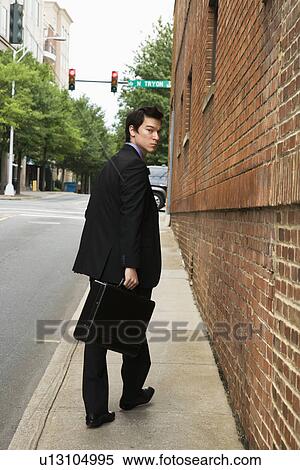 Stock Photography - Asian business man walking down sidewalk in the city looking back at viewer.. Fotosearch