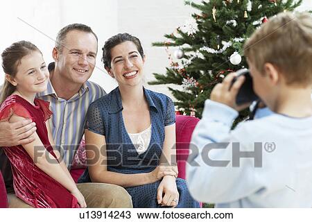 Stock Image - Boy taking photo of sitter mother and father by christmas tree. Fotosearch