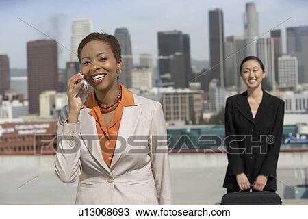 Stock Image - Business woman using mobile phone with another business woman behind in city. Fotosearch