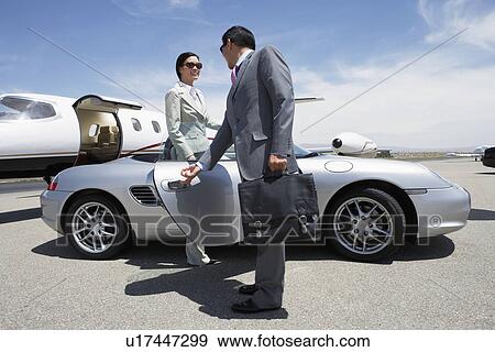 Stock Photo - Businessman holding door of convertible for colleague on landing strip near private jet. Fotosearch
