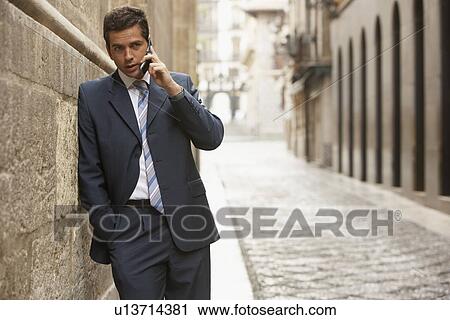 Stock Image - Businessman leaning on building in street talking on mobile phone.. Fotosearch