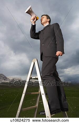 Stock Photography - Businessman on ladder in mountain field Speaking Through Megaphone low angle view. Fotosearch