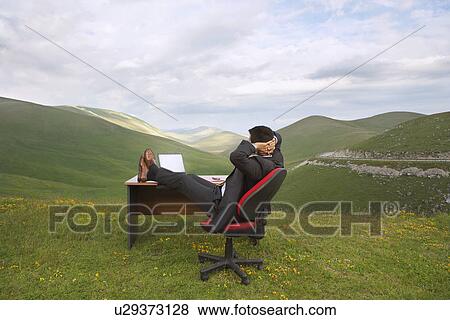 Businessman relaxing with feet on desk in mountain field side view View Large Photo Image Stock Photo - Businessman relaxing with feet on desk in mountain field side view. Fotosearch