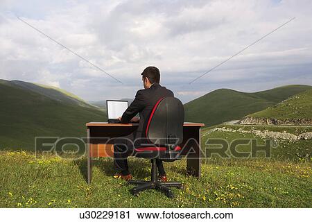 Businessman Sitting at Desk using laptop in mountain field back view View Large Photo Image Stock Image - Businessman Sitting at Desk using laptop in mountain field back view. Fotosearch