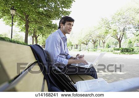 Stock Image - Businessman using laptop on park bench. Fotosearch