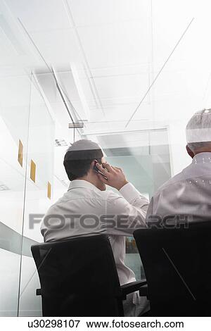 Businessmen one using mobile phone sitting in office behind glass wall back view View Large Photo Image Stock Photo - Businessmen one using mobile phone sitting in office behind glass wall back view. Fotosearch