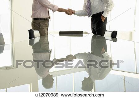 Stock Photo - Businessmen shaking hands by conference table mid section. Fotosearch