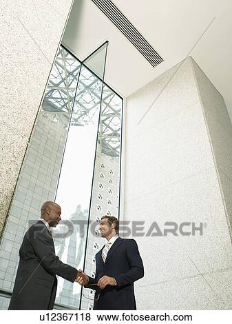 Stock Photo - Businessmen shaking hands in office building (low angle view). Fotosearch