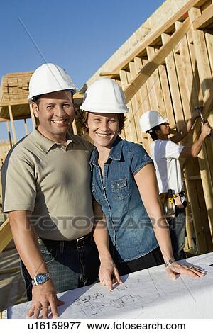 Couple in hard hats with blueprint on house construction site portrait View Large Photo Image Stock Photo - Couple in hard hats with blueprint on house construction site portrait. Fotosearch