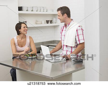 Stock Image - Couple in kitchen woman eating man using laptop. Fotosearch