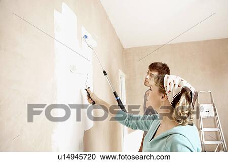 Stock Image - Couple painting wall with paint rollers indoors. Fotosearch