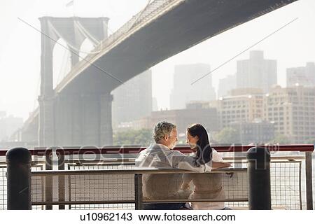 Stock Image - Couple relaxing on bench under Brooklyn Bridge. Fotosearch