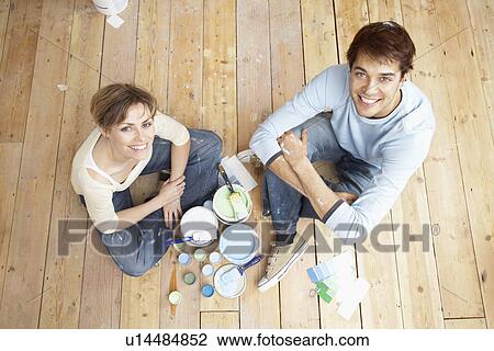 Stock Image - Couple sitting on floorboards amongst paint pots view from above. Fotosearch