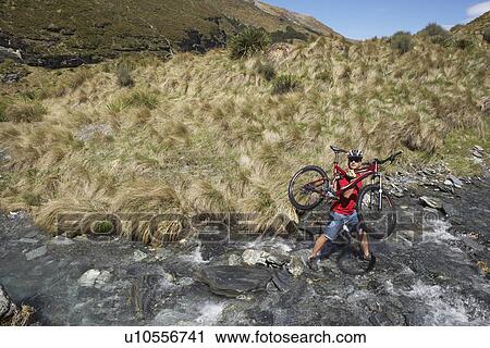 Stock Image - Cyclist carrying bike in river. Fotosearch