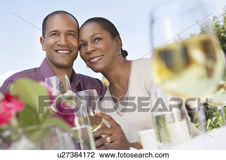 Stock Image - Elegant couple cheek to cheek holding wine glasses at garden party table. Fotosearch