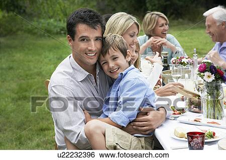 Family dining at table in garden father and son (7-9) looking at camera View Large Photo Image Stock Photograph - Family dining at table in garden father and son (7-9) looking at camera. Fotosearch