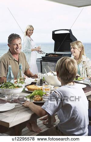 Family sitting around table outside eating Barbecue mother standing at grill grilling View Large Photo Image Stock Photo - Family sitting around table outside eating Barbecue mother standing at grill grilling. Fotosearch