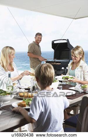 Family sitting around table outside eating Barbecue father standing at grill grilling View Large Photo Image Stock Photo - Family sitting around table outside eating Barbecue father standing at grill grilling. Fotosearch