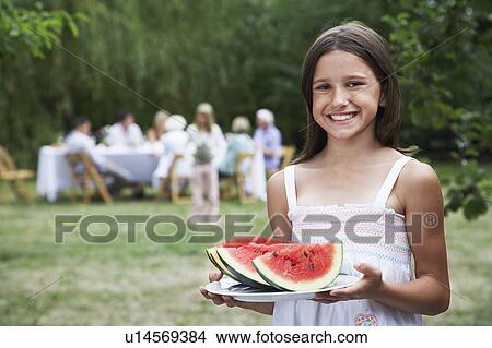 Girl (10-12) holding slices of watermelon in garden family members in background View Large Photo Image Picture - Girl (10-12) holding slices of watermelon in garden family members in background. Fotosearch