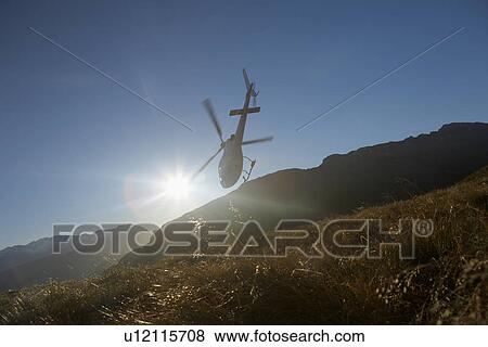 Stock Photo - Helicopter flying over hills in front of the sun. Fotosearch