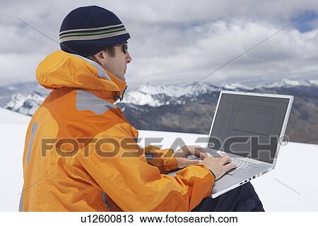 Stock Image - Hiker using laptop on snowy mountain peak. Fotosearch
