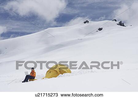 Stock Image - Hiker using laptop outside of tent on snowy mountain peak. Fotosearch