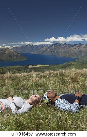 Stock Photo - Man and woman lying in field overlooking hills and lakes. Fotosearch