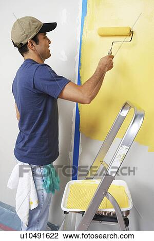 Stock Image - Man applying yellow paint to interior wall. Fotosearch