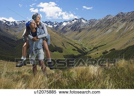 Stock Photography - Man giving woman piggy-back ride up hill with mountains behind. Fotosearch