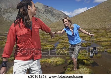 Stock Photo - Man helping woman wade through pond while she is carrying shoes. Fotosearch