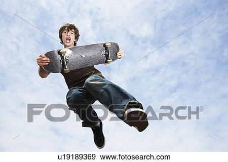 Man jumping holding skateboard in mid-air low angle view View Large Photo Image Stock Photo - Man jumping holding skateboard in mid-air low angle view. Fotosearch