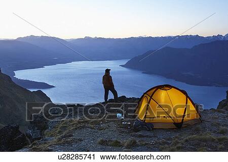 Man looking at lake next to tent at dusk View Large Photo Image Stock Image - Man looking at lake next to tent at dusk. Fotosearch