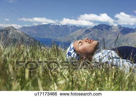 Stock Photograph - Man lying in field by lake and hills. Fotosearch