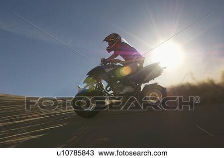 Stock Image - Man riding quad bike in desert against sun. Fotosearch