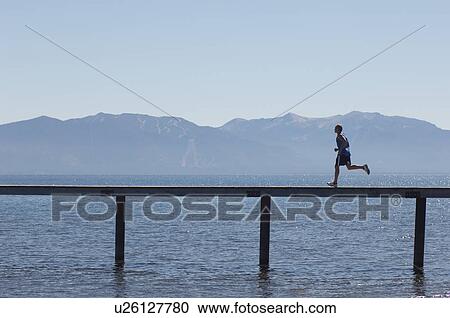 Stock Image - Man running along pier with mountains behind side view. Fotosearch