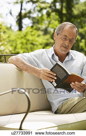 Stock Image - Man sitting on sofa in back yard reading book. Fotosearch