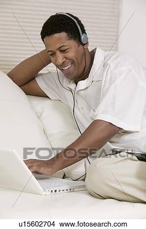 Man sitting on sofa using laptop with headphones View Large Photo Image Picture - Man sitting on sofa using laptop with headphones. Fotosearch