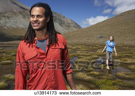 Stock Image - Man standing in field while woman wades in pond behind. Fotosearch