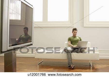 Stock Photograph - Man using laptop on modern sofa in modern apartment. Fotosearch