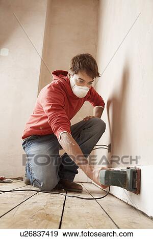 Stock Photo - Man wearing face mask sanding wall in unrenovated room low angle view. Fotosearch