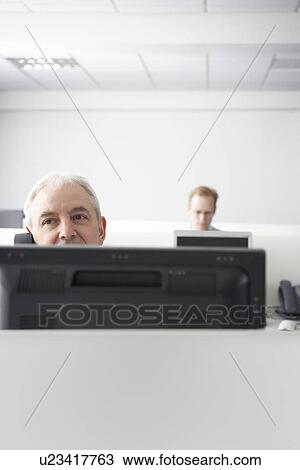 Stock Image - Mature businessman using telephone behind monitor in office cubicle high section. Fotosearch