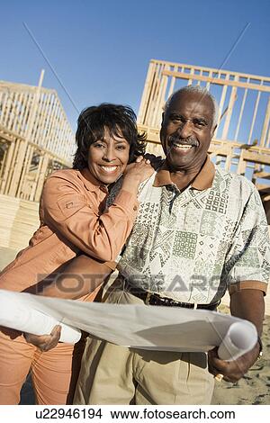 Picture - Middle-aged couple holding blueprint in front of house construction site portrait. Fotosearch