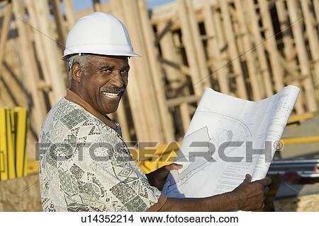 Middle-aged man in hard hat holding blueprint in front of house construction site View Large Photo Image Picture - Middle-aged man in hard hat holding blueprint in front of house construction site. Fotosearch
