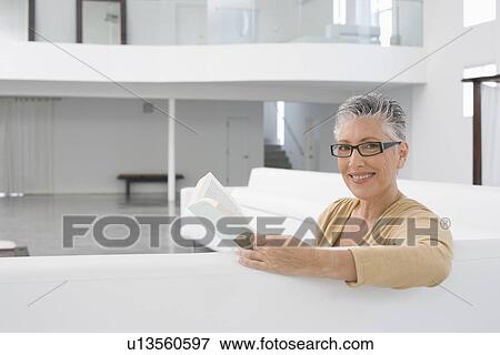 Stock Photo - Middle-aged woman reading book on sofa. Fotosearch