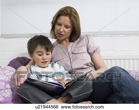 Stock Photograph - Mother and son (5-6) reading book on couch in living room. Fotosearch