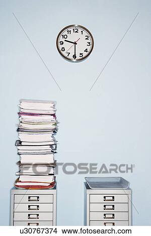 Picture - Office wall with clock stack of paperwork in inbox on file cabinet. Fotosearch