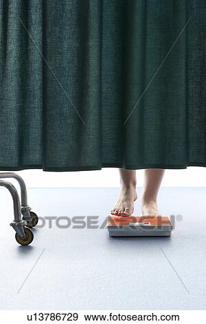 Stock Photo - Person stepping onto weighing scales behind curtain in hospital low section. Fotosearch