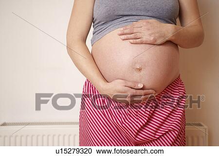 Stock Image - Pregnant woman holding exposed belly mid section. Fotosearch