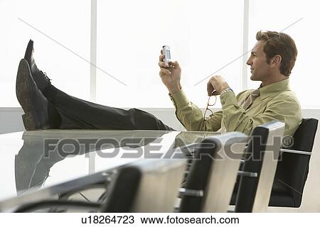 Stock Image - Relaxed businessman with feet up text messaging on mobile phone in conference room. Fotosearch