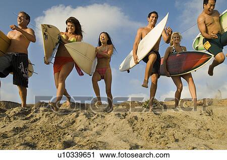 Stock Image - Six surfers carrying surfboards running on beach low angle view. Fotosearch